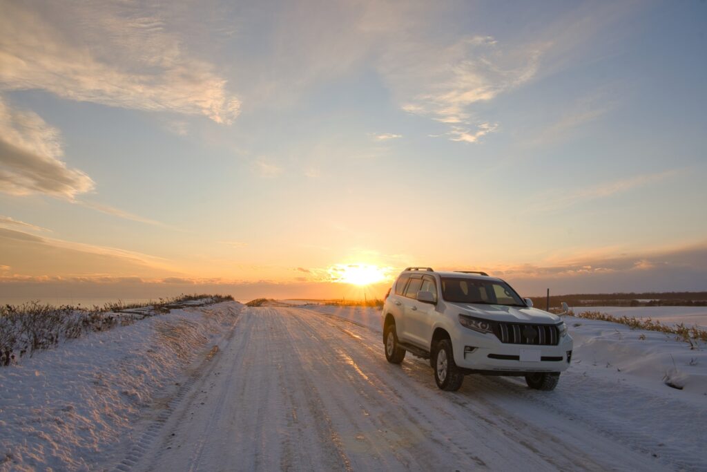 夕阳下，一輛白色SUV停在覆滿積雪和冰層的鄉村道路上，遠處是寬廣的地平線和金色陽光，突顯日本冬季自駕旅途的壯麗與安全感。