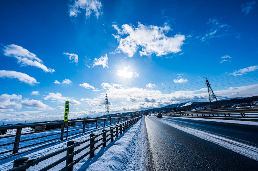 冬季晴朗的高速公路風景，路面上部分覆有雪痕，旁邊設有防護欄，遠處可見覆蓋白雪的山丘和電塔，藍天白雲襯托出冬季日本自駕的壯麗景色。